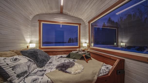 巨大なガラス窓のある木造キャビンの居心地の良いベッドルームの静的なビューは 時間経過とともに夜明けに雪に覆われた田舎を外に見る — ストック動画