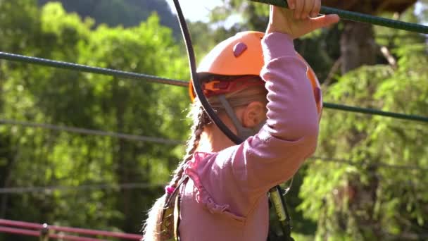 両方の手でロープを保持しながら クライミングパーク内の課題を探索する女の子 ヘルメットと森の中で安全ハーネスを持つ6歳のヨーロッパの女の子 浅い深さの背景とスローモーション — ストック動画