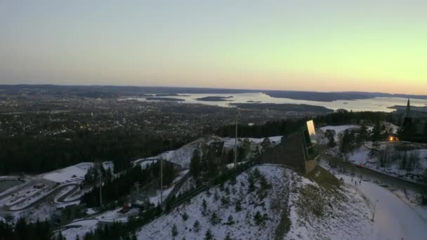 オスロの街並みドローンは Holmenkolbakken Vinterpark Winterpark Tryvann 前景にHolmenkollスキージャンプでドリーショットサンセットで過去のスキージャンプ — ストック動画