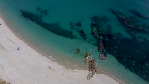 プラヤデルピノの外の透明な水の中の岩の上で波がクラッシュします 白い砂浜に2つの青の太陽の傘 スペインの南海岸のほぼ空のビーチ — ストック動画