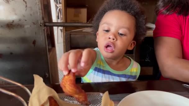 エキゾチックな2歳のアフリカ系ヨーロッパ人の子供せっかちで興奮した彼の母親の隣に座っているレストラン内のホットチキンストリップを食べる — ストック動画