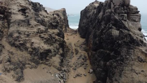岩の多いビーチで山を歩く人のドローンビデオ ドローンは峡谷に飛び込み 海に向かって人を飛び越える ペルーのリマにあるチョリロスに位置します ビーチ — ストック動画