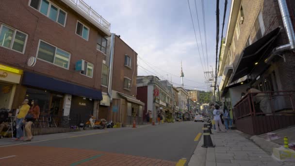 数多くの西洋レストランやお店が立ち並ぶ現代的な龍山海番村通りと日没時のランドマーク的なNソウル南山タワーの景色 ワイド確立ショット — ストック動画