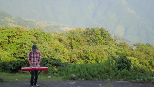 男の静止した映像 背景に山の広い角度ビューで彼の電子音楽キーボードを再生 彼はまた 曲を変更するために彼のキーボードに接続されたペダルを使用します — ストック動画