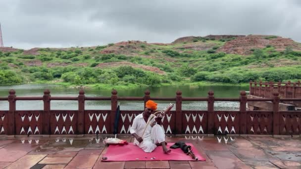牧歌的な風景の中で湖の前で伝統的な楽器を演奏座って地元のインド人の男の静的なショット — ストック動画