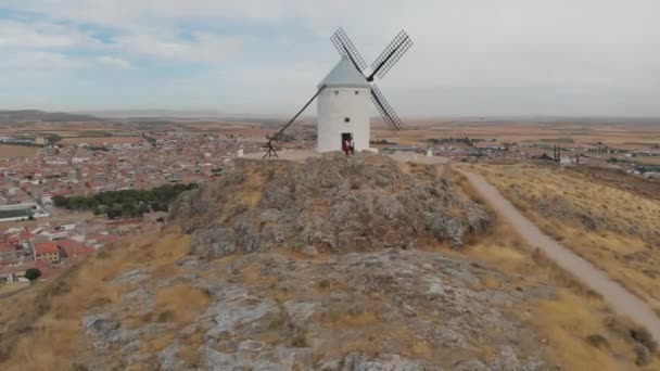 丘の上の孤独な古い白い風車の前に立っている2人の観光客からのプルバック空中ショット — ストック動画