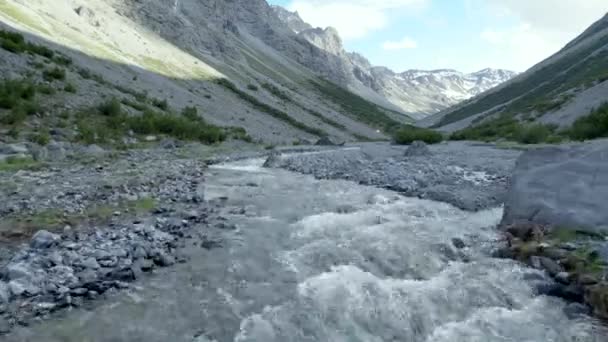 スイスに雪が残る急峻で劇的な山の風景に囲まれた氷河の谷の流れの速い川の近くを飛んでいる空中ドローン映像 — ストック動画