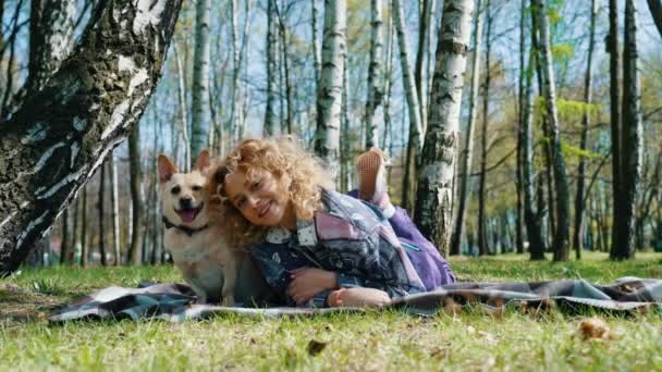 公園でカメラを見ながら笑顔で胃の上に寝そべっている少女の横に座っているペンブロークウェールズのコーギー犬 — ストック動画