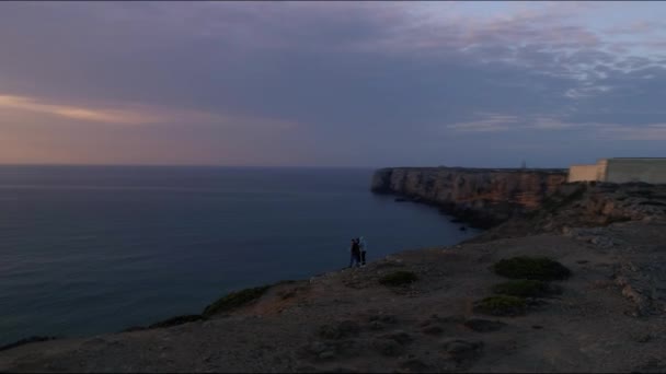 ポルトガルのサグレス砦のオレンジ色の青い日の出の夕日を見て崖の端に立っている3人 周囲の空中でドローンが撃たれ — ストック動画