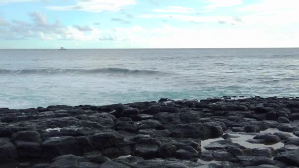 Hdスローモーションハワイカウアイ島の船はフレームの左端から右に向かって左に進み 海の波が前に溶岩の岩があり 部分的に曇っている空のカメラに向かって建物 — ストック動画