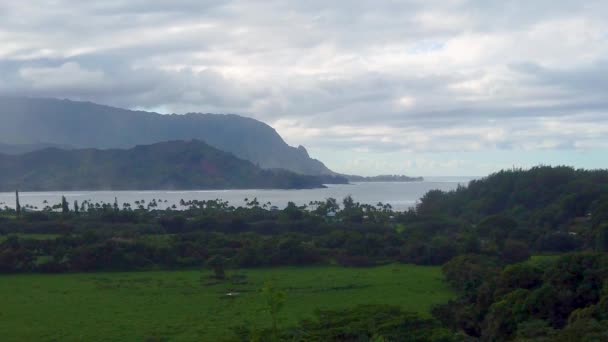 Hawaii Kauaiゆっくりとした動きほとんど曇りの空との距離で入り江や山岳海岸線と草原フィールドの静的なワイドショット — ストック動画