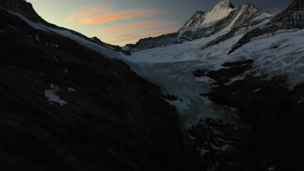 航空写真は 暗い谷と影の中の氷の氷河と日の出または日没でオレンジ 黄色と青のカラフルな空のゆっくりと傾斜します スイスのグレックシュタイン ハットで — ストック動画