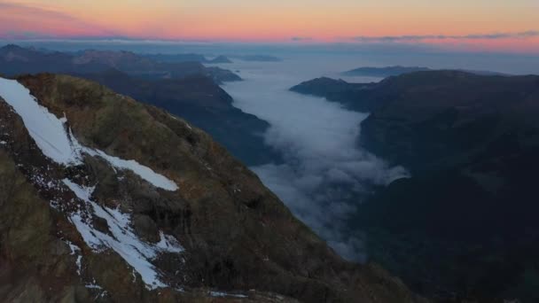 慢腾腾的无人机拍摄 暴露了冰雪覆盖的光秃秃而寒冷的山顶 下面是一个巨大的山谷 云雾笼罩着它 瑞士夏天多姿多彩美丽的日出 — 图库视频影像