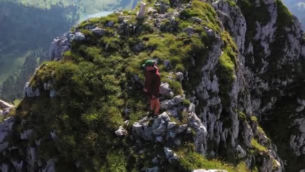 パーカーとキャップを着た若い男がピークの端に立っていて 驚くほど劇的な風景を見ています スイスアルプスのストックホルムの茂みや岩に囲まれて — ストック動画
