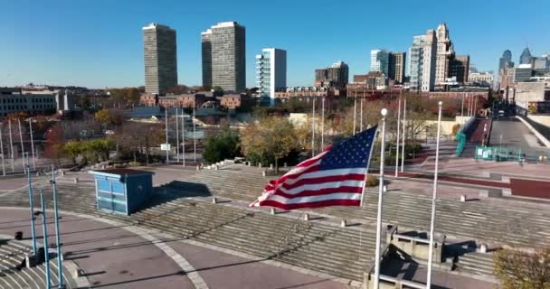 風の中でアメリカの旗の波 遠くの都市アメリカの都市 静かな街の風景 スローモーションでの空中ドローンの軌道 — ストック動画