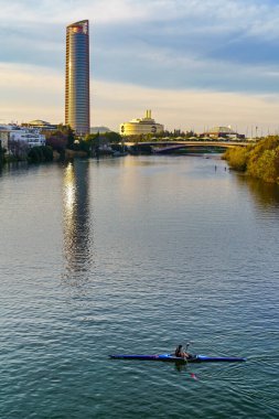 Seville 'deki Guadalquivir nehrinde gün batımı manzarası nehirde kürek çeken tekneler ve sudaki binaların yansımaları. İspanya.