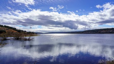Göl manzarası, sudaki bitkiler ve ağaçlar, karanlık bulutlar ve sudaki güneşin yansımaları. Madrid Guadalix. Avrupa.