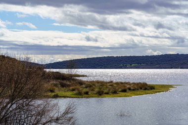 Göl manzarası, sudaki bitkiler ve ağaçlar, karanlık bulutlar ve sudaki güneşin yansımaları. Madrid Guadalix. Avrupa.