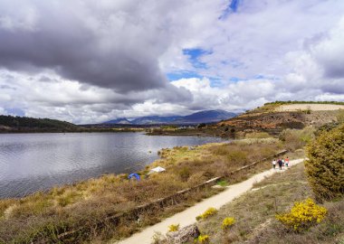 İnsanlar mavi göl kıyısında su ve fırtına bulutlarındaki yansımalarla geziniyorlar. Guadalix Madrid.