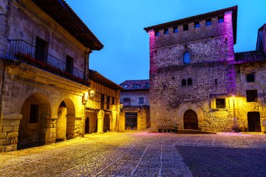 Kale kulesi ve kemerli kapıları olan eski kasaba meydanı, geceleri aydınlık. Santillana del Mar.