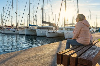 Önünde güneş olan bir bankta dinlenen kadın limandaki teknelere bakıyor..