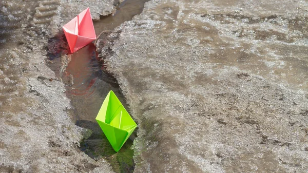 Green and pink paper boats float on the stream on the road in spring.