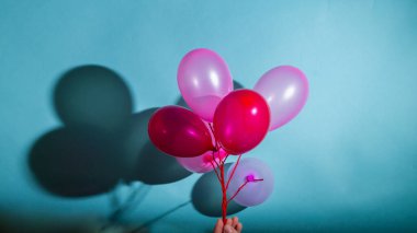 Bir adam elinde mavi arka planda bir demet pembe ve kırmızı balon tutuyor..