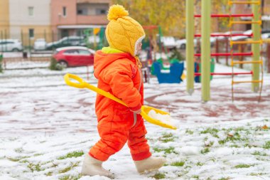 Turuncu tulumlu bir kız bahçede kürekle karla oynuyor..