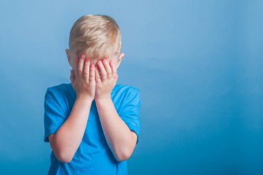 Şaşırma duygusu. Mavi tişörtlü bir çocuk ellerini stüdyonun mavi arka planına sürerek yüzünü kapattı..