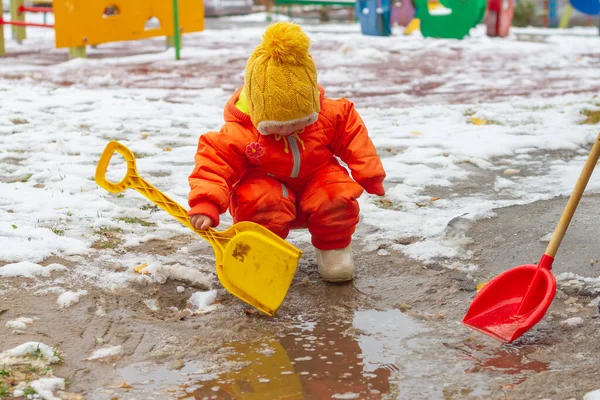 Turuncu tulumlu bir kız bahçede kürekle karla oynuyor..