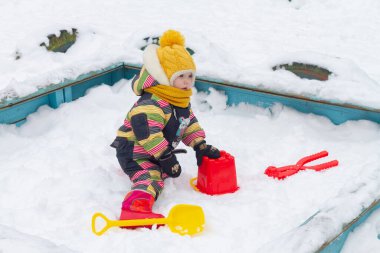 Kışın evin avlusunda kırmızı bir kova ve sarı bir kürekle oynayan bir kız çocuğu kovaya plastik kar döker..
