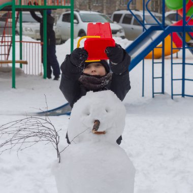 A preschool boy made a snowman out of snow in winter and puts a red bucket on the snowman's head.