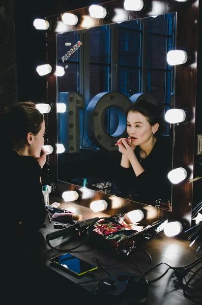 Giovane bella donna che applica le labbra make-up, guardando in uno specchio, seduta sulla sedia a teatro spogliatoio con specchio d'epoca con lampadine camera oscura Foto Stock