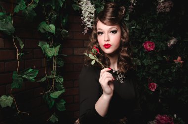 Dalgalı saçlı, kırmızı dudaklar, siyah elbiseli, romantik güzel kız gül çiçek çiçek açan bahçede holding sihirli portresi. Moda kadın peri masalı Prenses, mistery orman yürüyüş hakkında. Yaratıcı sanat 