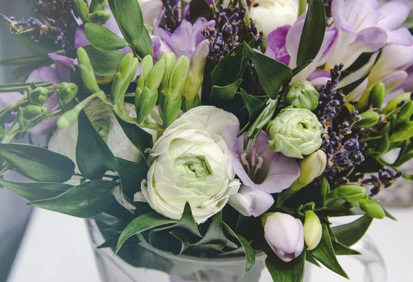 Ricco mazzo di ranuncolo bianco e fresia lilla, fiori di rose, foglia verde in vaso di vetro. Bouquet fresco primavera estate. Moda sfondo floreale . — Foto Stock