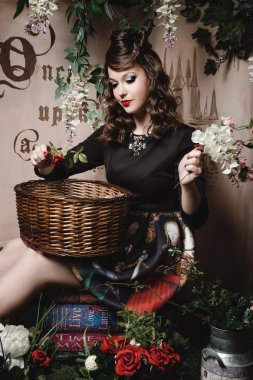 Sanat kitapları üzerinde oturan ve bahar çiçekleri sepeti ile poz güzel genç esmer kadın. Sihirli portre romantik kız dalgalı saçlı, kırmızı dudaklar, gül tutan elbise, moda. Erkek sahne