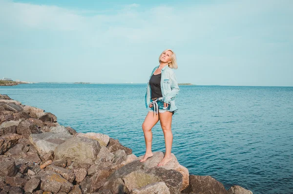 Eine braun gebrannte Frau posiert an einem Sommertag am Strand und blickt auf. verträumte Frau in Jeans-Klamotten, felsige Küste stehend, blauer Himmel mit Meer. Konzepturlaub im Freien. Reiseaktiver Lebensstil. — Stockfoto