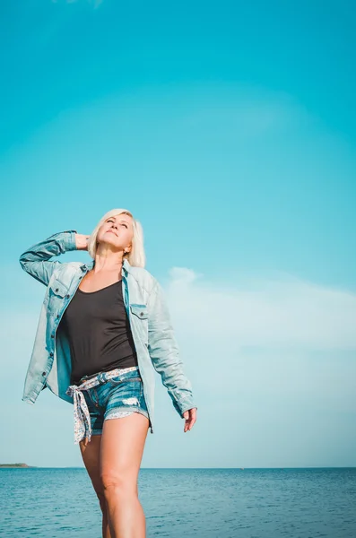 Tanned woman posing at the beach on summer day, looking up. Dreamy female in jeans clothes, standing rocky coast, blue sky background with sea. Concept holiday, outdoors. Travel active lifestyle.