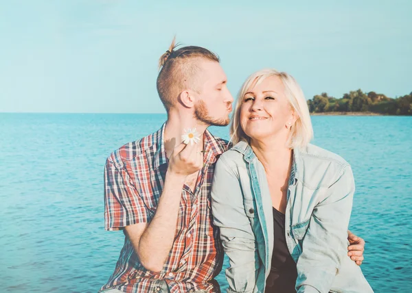 Il figlio bacia e abbraccia sua madre nel giorno d'estate, all'aperto. Madre il suo bambino adulto divertirsi insieme. Il concetto di madre. Felicità famiglia seduta sulla spiaggia rocciosa con acqua blu, sfondo cielo. Amore. Fotografia Stock