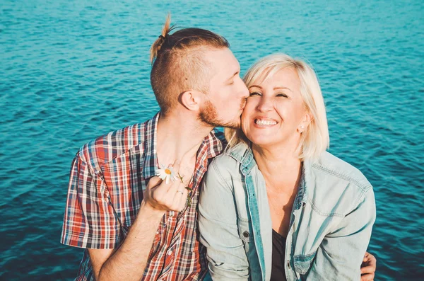 Il figlio bacia e abbraccia sua madre nel giorno d'estate, all'aperto. Madre il suo bambino adulto divertirsi insieme. Il concetto di madre. Felicità famiglia seduta sulla spiaggia rocciosa con acqua blu, sfondo cielo. Amore. Immagini Stock Royalty Free