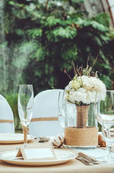 Conjunto de mesa para una fiesta o banquete de recepción de boda. Ramo nupcial de invierno de claveles blancos, peonía, rosas flores con cordel en jarrón sobre fondo artesanal. Elegante copa de vino, cubiertos, servilleta —  Fotos de Stock