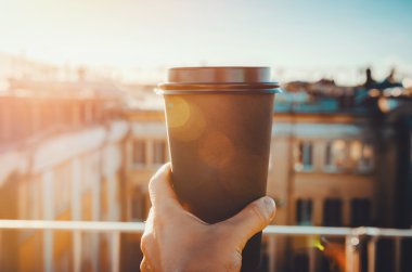 hands holding hot craft cup of coffee or tea in morning sunlight with view to blurred city background. Enjoy, lifestyle, take away breakfast concept. woman on the roof   drink clipart
