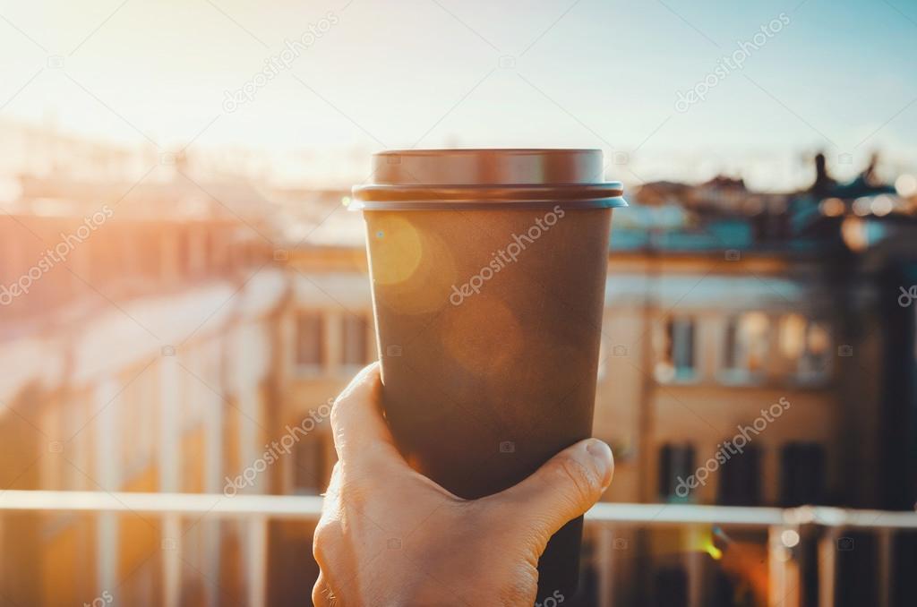 hands holding hot craft cup of coffee or tea in morning sunlight with view to blurred city background. Enjoy, lifestyle, take away breakfast concept. woman on the roof   drink