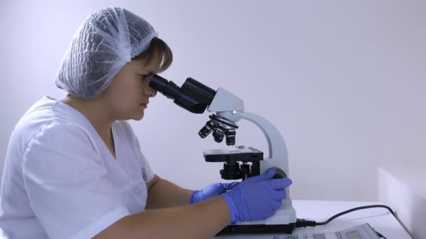 Woman laboratory technician comes and sit down at his workplace under microscope — Stock Video