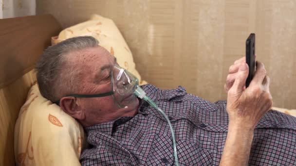 A senior man in oxygen mask is lying on bed and watching something in his smartphone — Stock Video