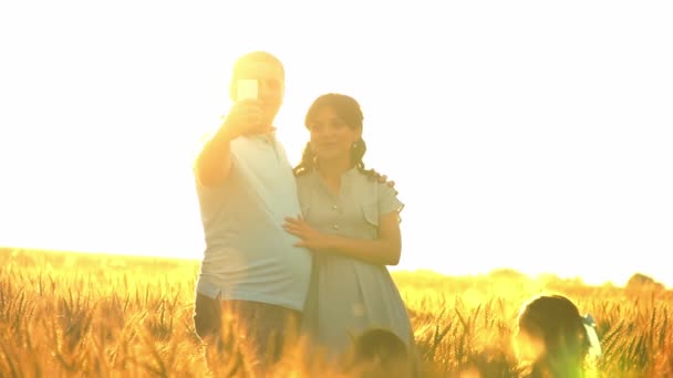 Una pareja está haciendo selfie al atardecer en un campo de trigo con dos niños en cámara lenta — Vídeos de Stock