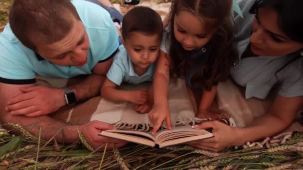 Szczęśliwa para z dwójką dzieci kłamie i czyta książkę na łące w slowmo — Wideo stockowe