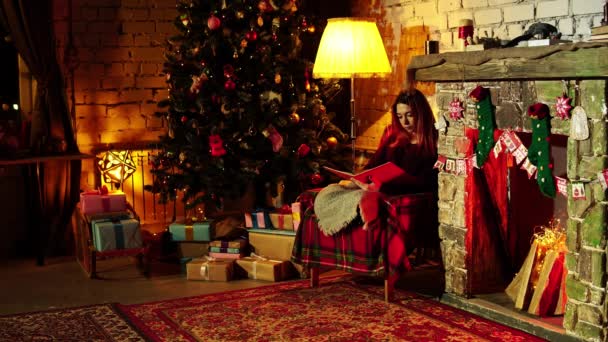 女性は美しい装飾されたクリスマスツリーを見て楽しむと本とアームチェアに残ります — ストック動画