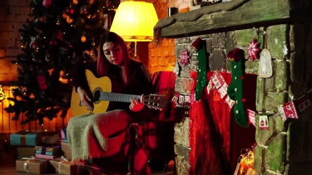 Mujer se sienta cerca del árbol de Navidad y toca la guitarra — Vídeos de Stock