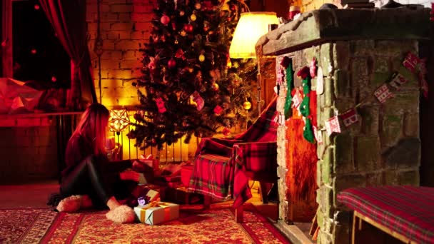 A young woman is hiding gifts under a fir at Christmas Eve sitting in a festive decorated living room — Stock Video